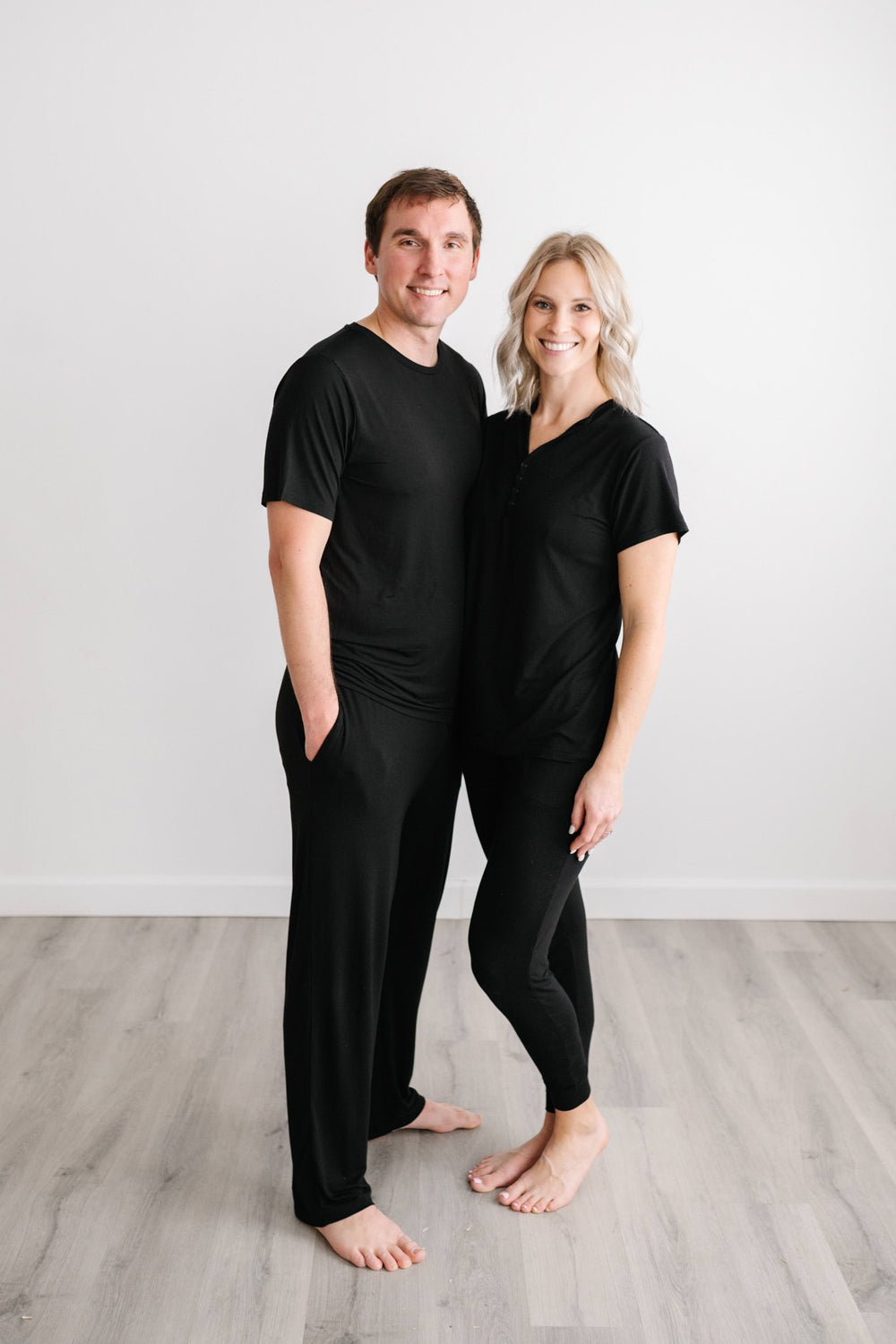 Image of a couple wearing matching solid black pajamas. They are both shown wearing short sleeve pajama tops with matching pajama bottoms. 