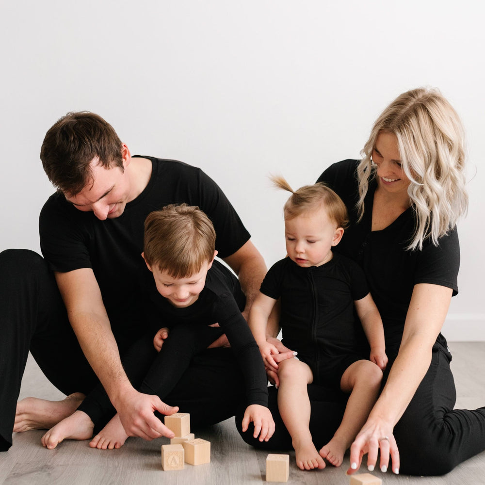 Click to see full screen - Image of family of 4 all sitting down on the floor and playing with wooden building blocks. They are all shown wearing matching solid black pajamas. The mom and dad are both shown wearing solid black short sleeve pj tops with matching solid black pj pants
