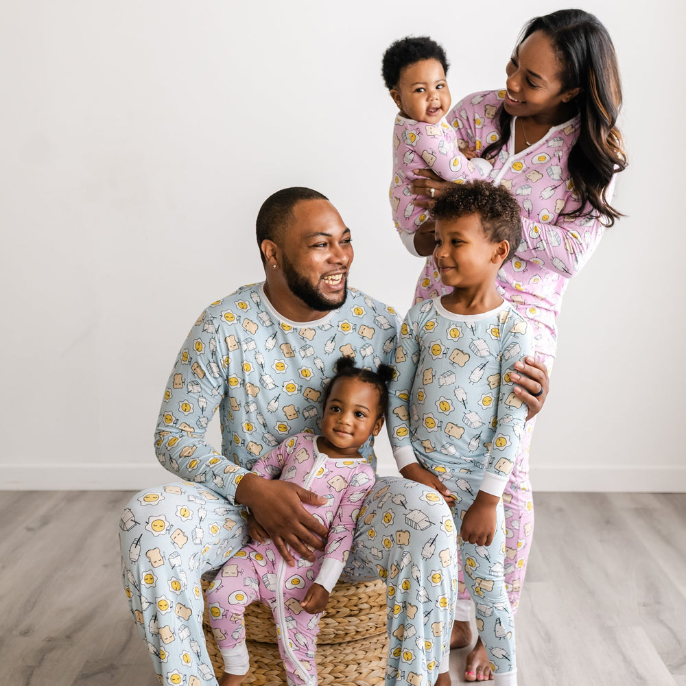 Image of family of five all wearing matching Breakfast Buddies printed pajamas. Father and son are wearing Blue Breakfast Buddies printed pajamas with white trim accents, while mom is shown wearing Pink Breakfast Buddies printed pajamas with white trim accents. The two daughters in the photo are also wearing Pink Breakfast Buddies printed Zippies with white trim accents. The breakfast foods featured on this print include sunny side up eggs, toast, and milk.