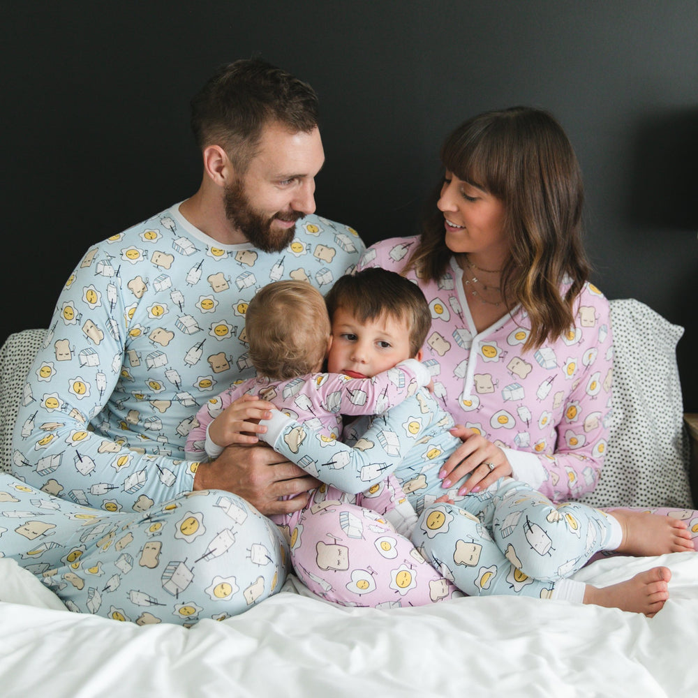 Image of family of four all wearing matching Breakfast Buddies printed pajamas. Father and son are wearing Blue Breakfast Buddies printed pajamas with white trim accents, while mom and daughter are shown wearing Pink Breakfast Buddies printed pajamas with white trim accents. The breakfast foods featured on this print include sunny side up eggs, toast, and milk.