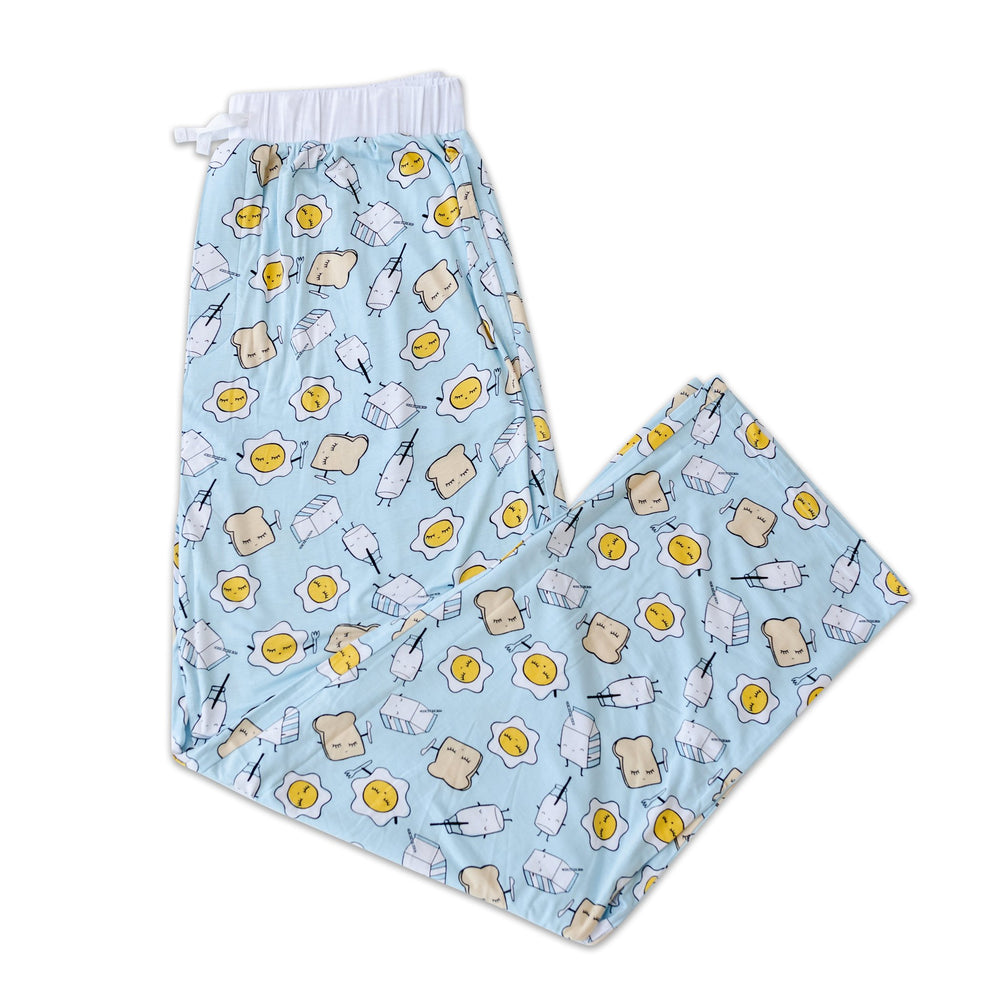 Flay lay photo of men's Blue Breakfast Buddies printed pajama pants. The pants are featured having an adjustable waist drawstring and pockets. The waist has a white trim accent, and the  breakfast foods featured on this print include sunny side up eggs, toast, and milk.