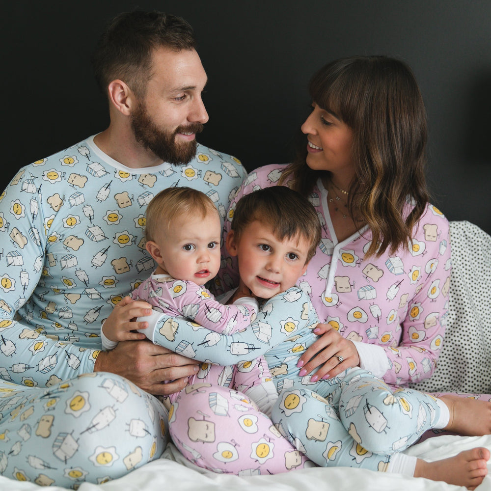 Click to see full screen - Image of family of four all wearing matching Breakfast Buddies printed pajamas. Father and son are wearing Blue Breakfast Buddies printed pajamas with white trim accents, while mom and daughter are shown wearing Pink Breakfast Buddies printed pajamas with white trim accents. The breakfast foods featured on this print include sunny side up eggs, toast, and milk.