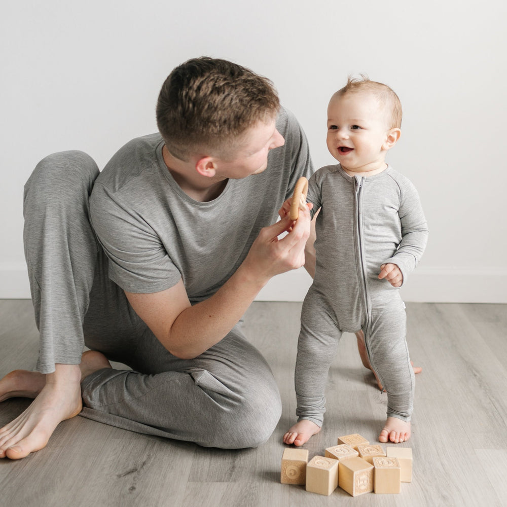 Image of a dad and son playing with wooden building blocks. The dad is shown wearing a short sleeve pajama top and pajama pants in heather gray, while the son is shown wearing a zip up romper in heather gray.