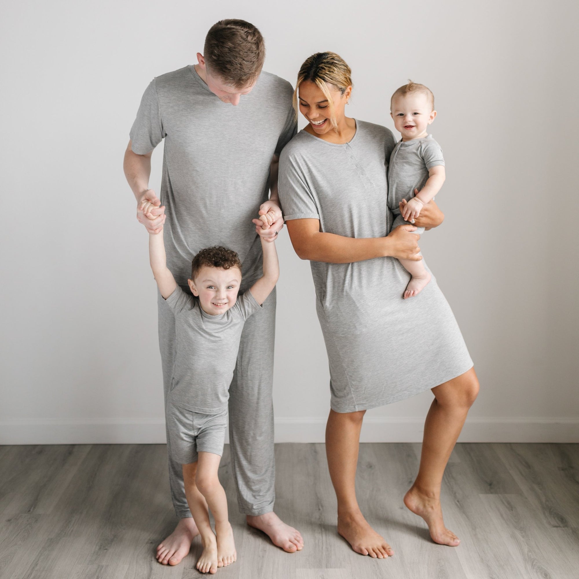 Image of family of four shown wearing matching solid heather gray pajamas. The dad is shown wearing a short sleeve pj top and pants, the mom is shown wearing a short sleeve caftan gown, one son is shown wearing a short sleeve and shorts pj set, while the other son is shown wearing a short sleeve and shorts zip up romper.