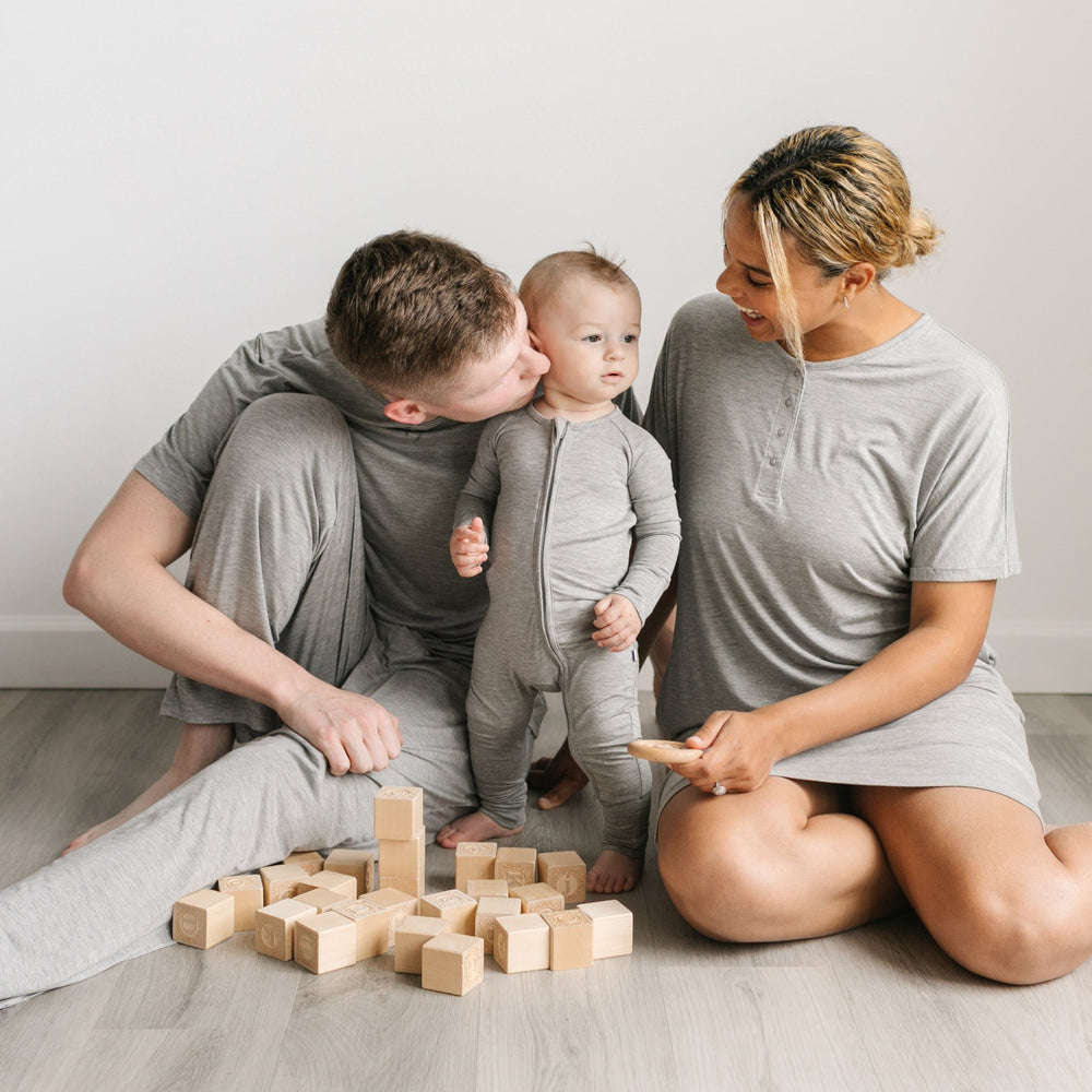 Image of a mom and dad playing with wooden building blocks with their infant son. They are all three shown wearing matching heather gray pajamas. The mom is shown wearing a short sleeve caftan gown, the dad is shown wearing a short sleeve pajama top with matching pajama bottoms, and the son is shown wearing a zip up romper.