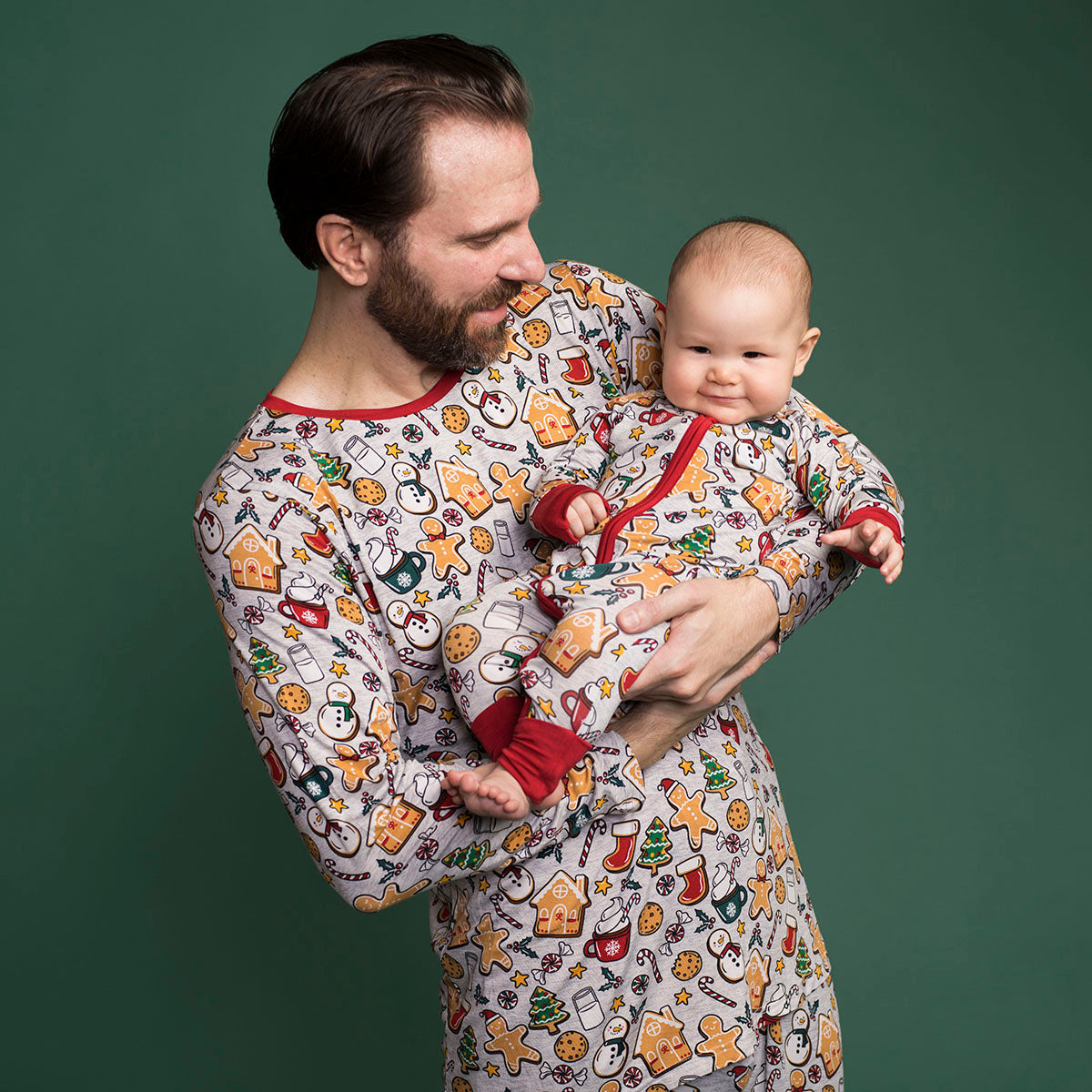 Father holding his son matching in Holiday Treats pajamas. Dad is wearing Holiday Treats men's pajama top paired with matching men's pajama pants. Child is wearing Holiday Treats zippy 