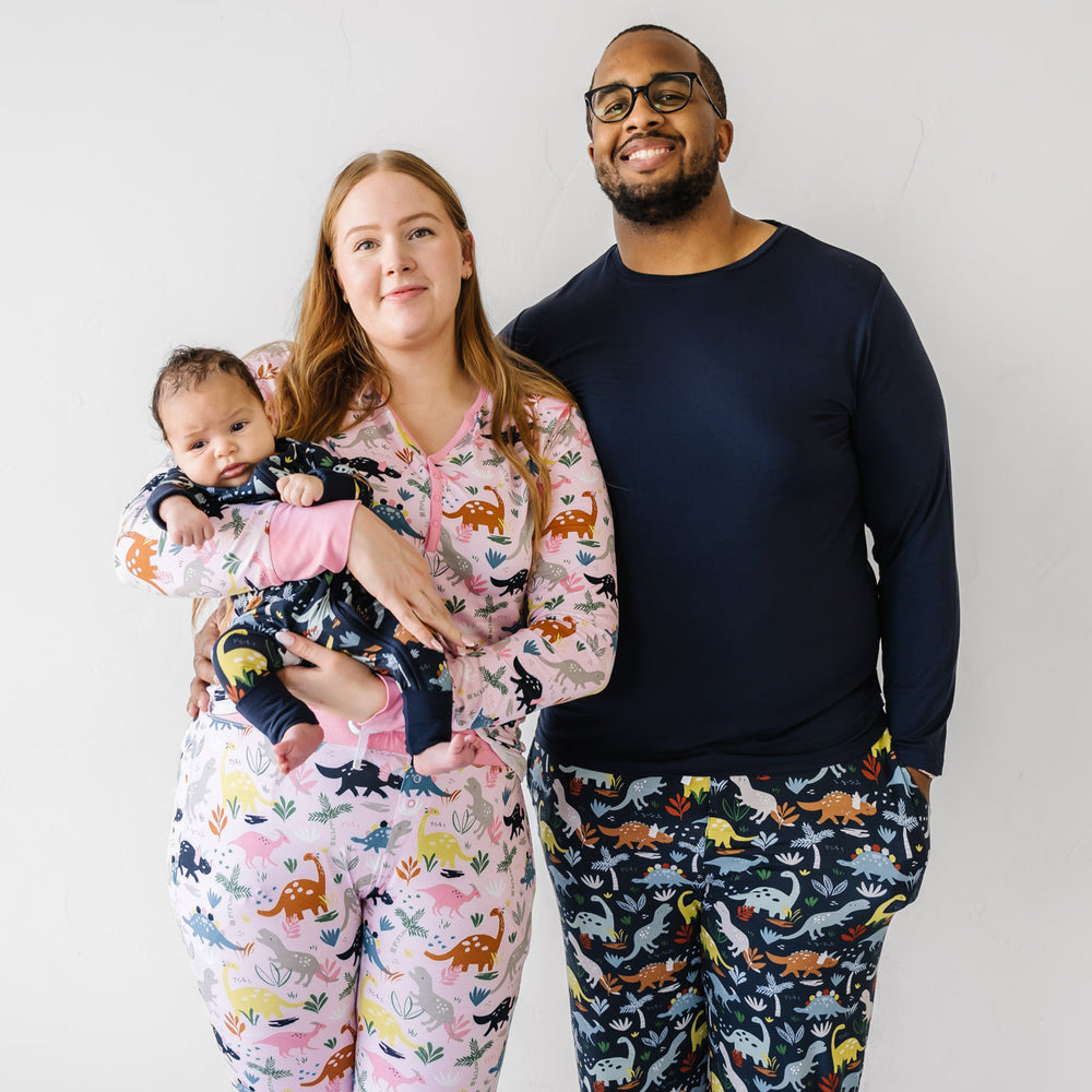 Family of thee posing together. Mother is wearing a women's Pink Jurassic Jungle printed pajama top paired with matching bottoms and father is wearing a coordinating men's Navy Jurassic Jungle pajama bottoms paired with a solid Navy top. Baby is matching wearing a Navy Jurassic Jungle printed zippy