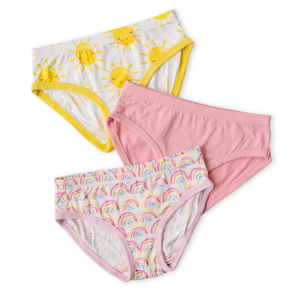 Scattered flat lay image of 3 sets of girls brief underwear. One yellow smiling sunshine printed style, followed by one solid bubblegum pink style, followed by one pastel pink rainbows printed style.