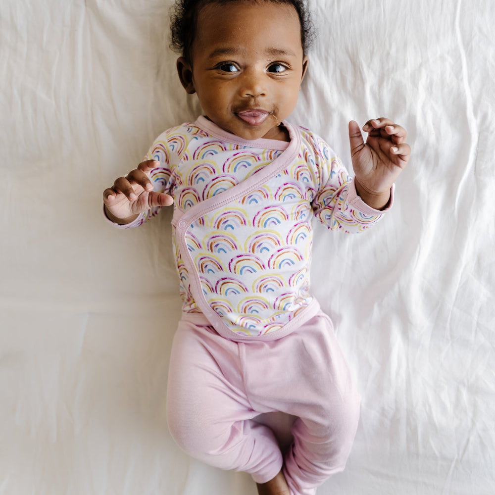 Image of an infant wearing a two-piece crossover set that features a long sleeve wrap style top with snap closures and coordinating pants with convertible footies. This style is in the Pastel Rainbows print that features watercolor style rainbows in a sweet pastel hues on a white background with soft pink trim.