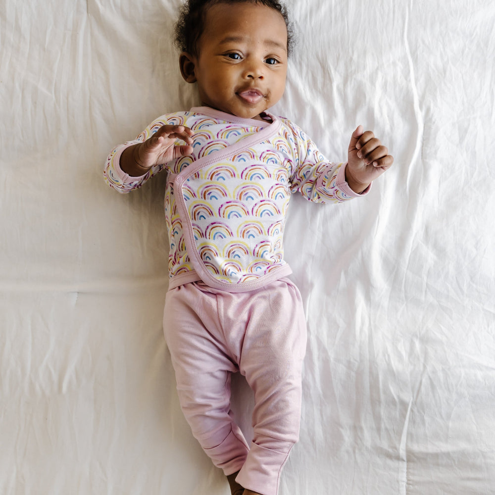 Image of an infant wearing a two-piece crossover set that features a long sleeve wrap style top with snap closures and coordinating pants with convertible footies. This style is in the Pastel Rainbows print that features watercolor style rainbows in a sweet pastel hues on a white background with soft pink trim.