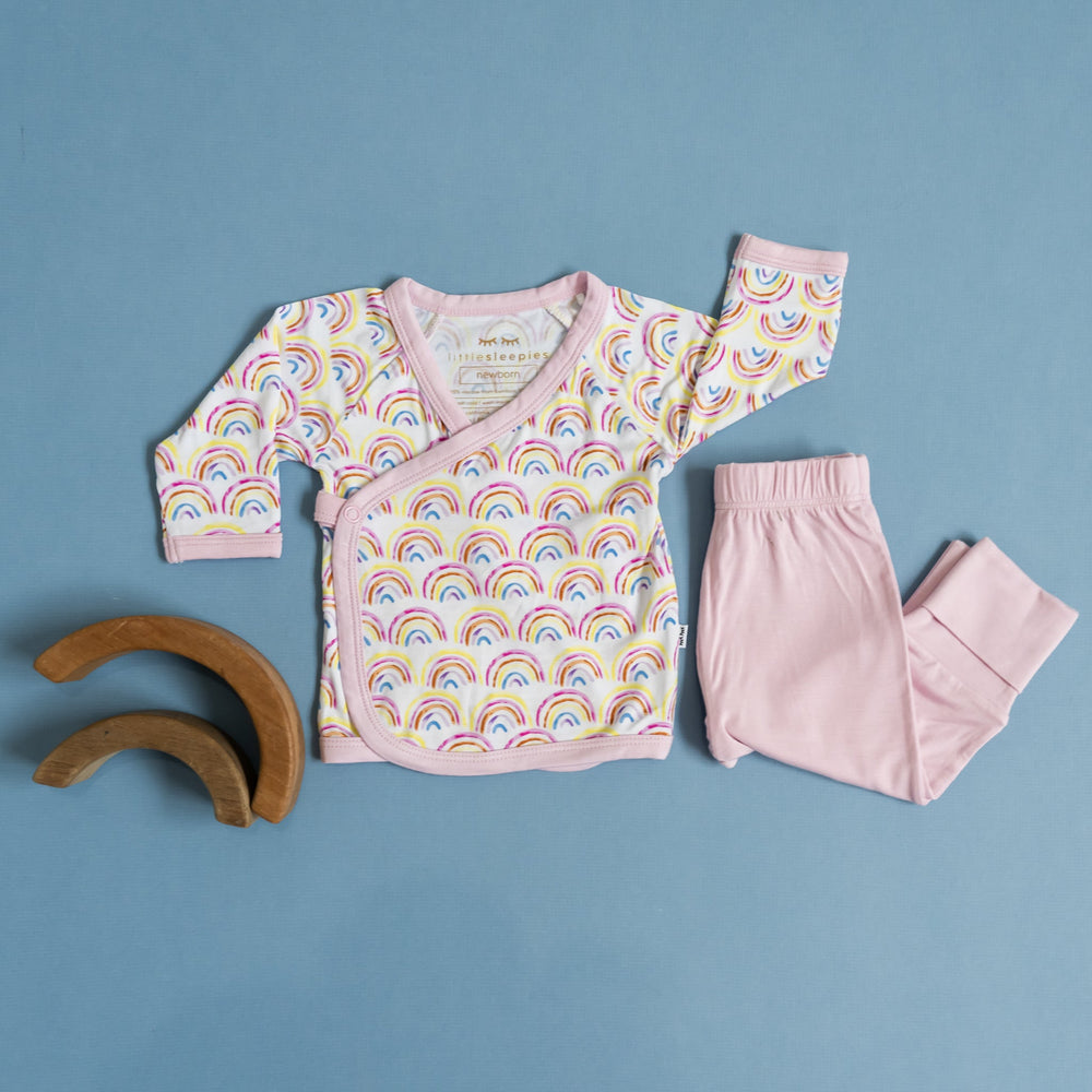 Flat lay image of a two-piece crossover set that features a long sleeve wrap style top with snap closures and coordinating pants with convertible footies. This style pictured has the wrap style top in Pastel Rainbows print with watercolor style rainbows in pastel hues on a white background with soft pink trim.