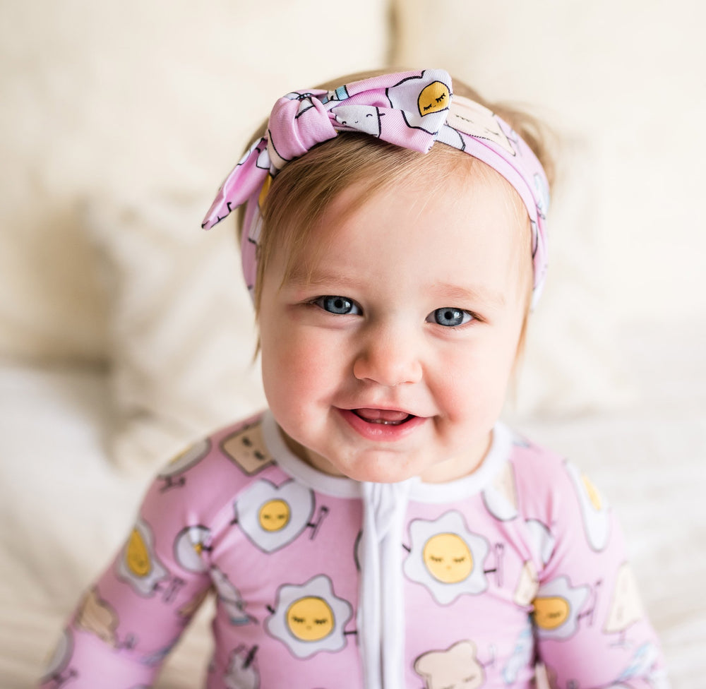 Click to see full screen - Image of infant girl wearing Pink Breakfast Buddies printed zip up romper with matching bow headband. This print has a light pink background with white trim accents and the breakfast foods featured on this print include sunny side up eggs, toast, and milk.