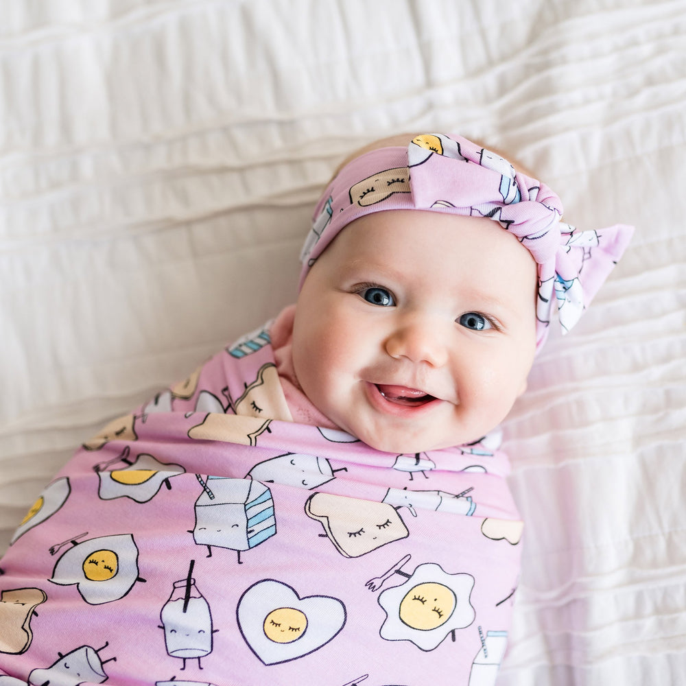 Image of infant girl wearing swaddle and headband set in Pink Breakfast Buddies print. 