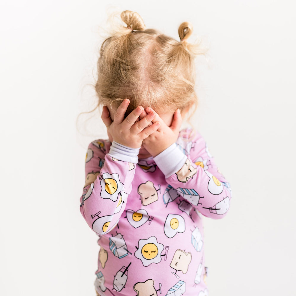 Image of toddler girl wearing two-piece pajama set in Pink Breakfast Buddies print. This print has a light pink background with white trim accents and the breakfast foods featured on this print include sunny side up eggs, toast, and milk.