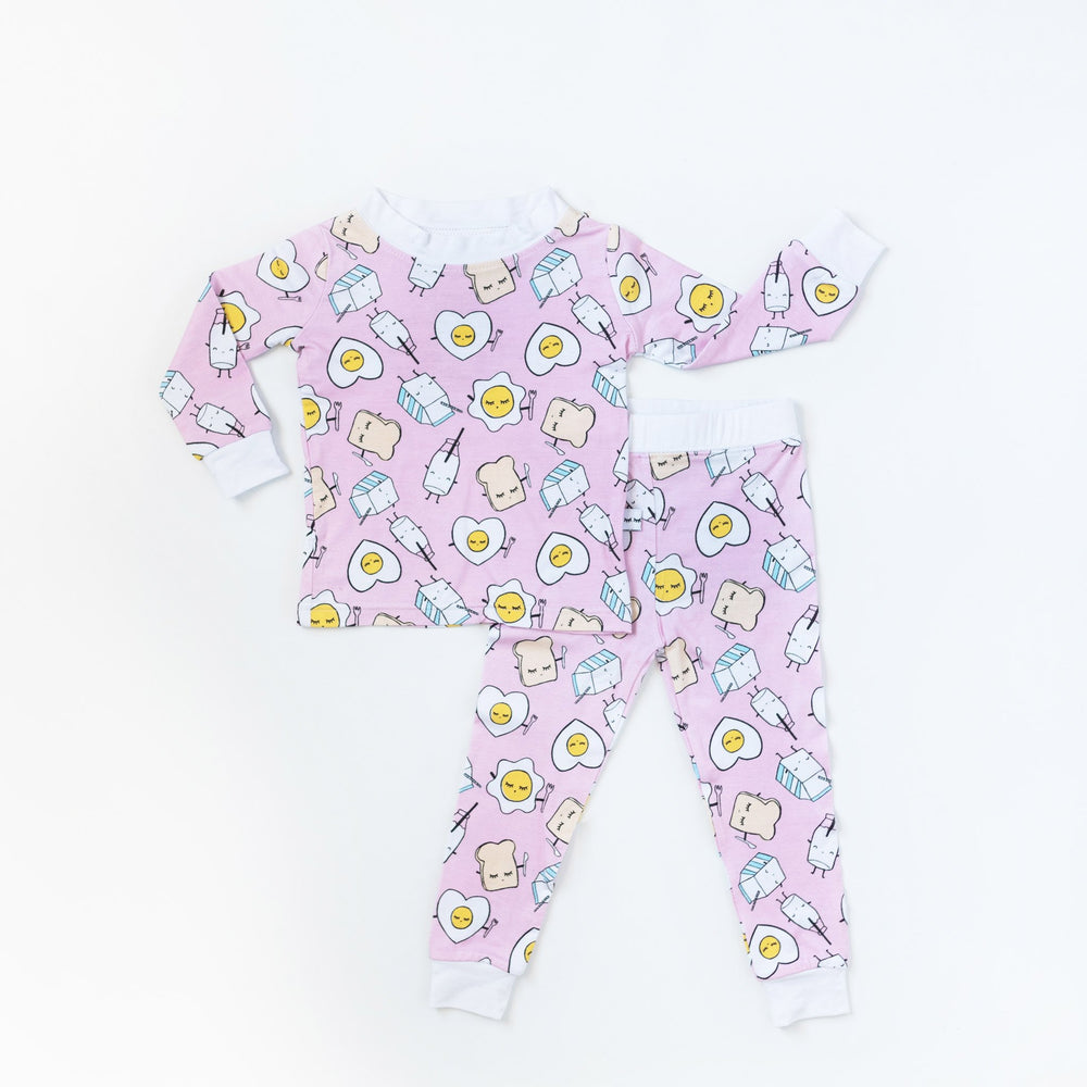 Flat lay image of two-piece pajama set in Pink Breakfast Buddies print. This print has a light pink background with white trim accents and the breakfast foods featured on this print include sunny side up eggs, toast, and milk.