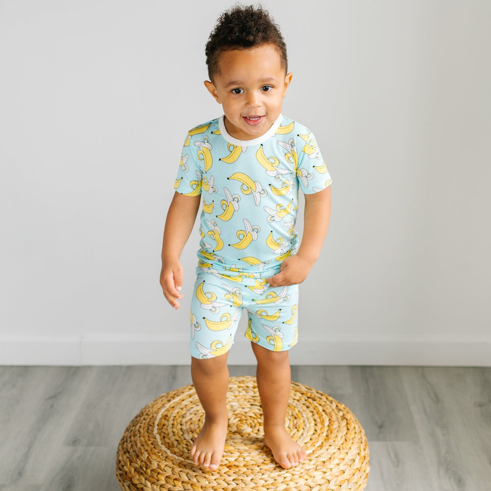 Image of little boy wearing a banana printed two-piece short sleeve and shorts pajama set. This print features a pop of bright yellow bananas that sit upon a blue background with white trim accents. 