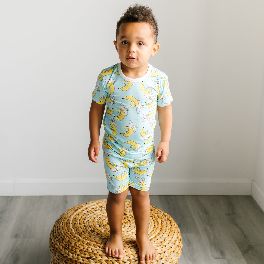 Image of little boy wearing a banana printed two-piece short sleeve and shorts pajama set. This print features a pop of bright yellow bananas that sit upon a blue background with white trim accents.