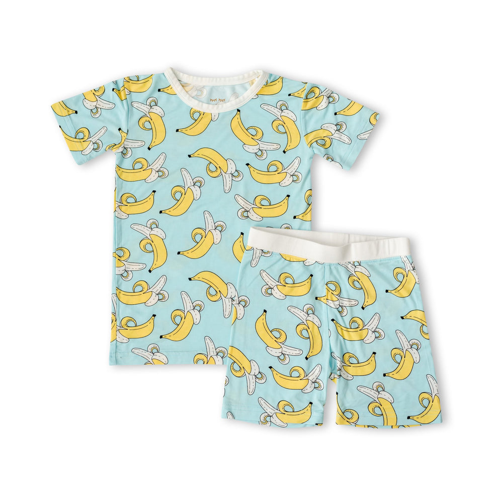 Flat lay image of banana printed two-piece short sleeve and shorts pajama set. This print features a pop of bright yellow bananas that sit upon a blue background with white trim accents. 