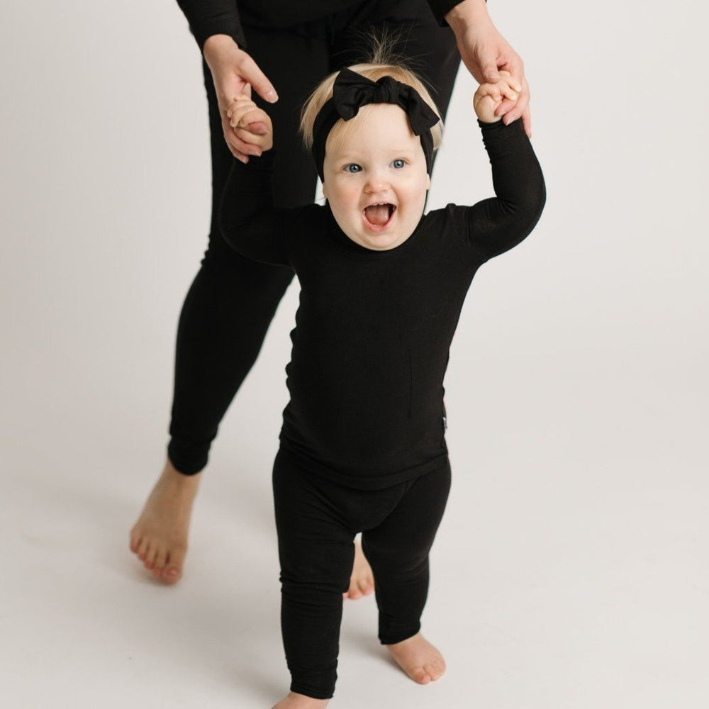 Baby girl wearing solid black two-piece pajama set with matching solid black bow headband. 