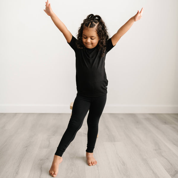 Image of little girl posing with her arms in the air. She is shown wearing a solid black short sleeve pajama set. 