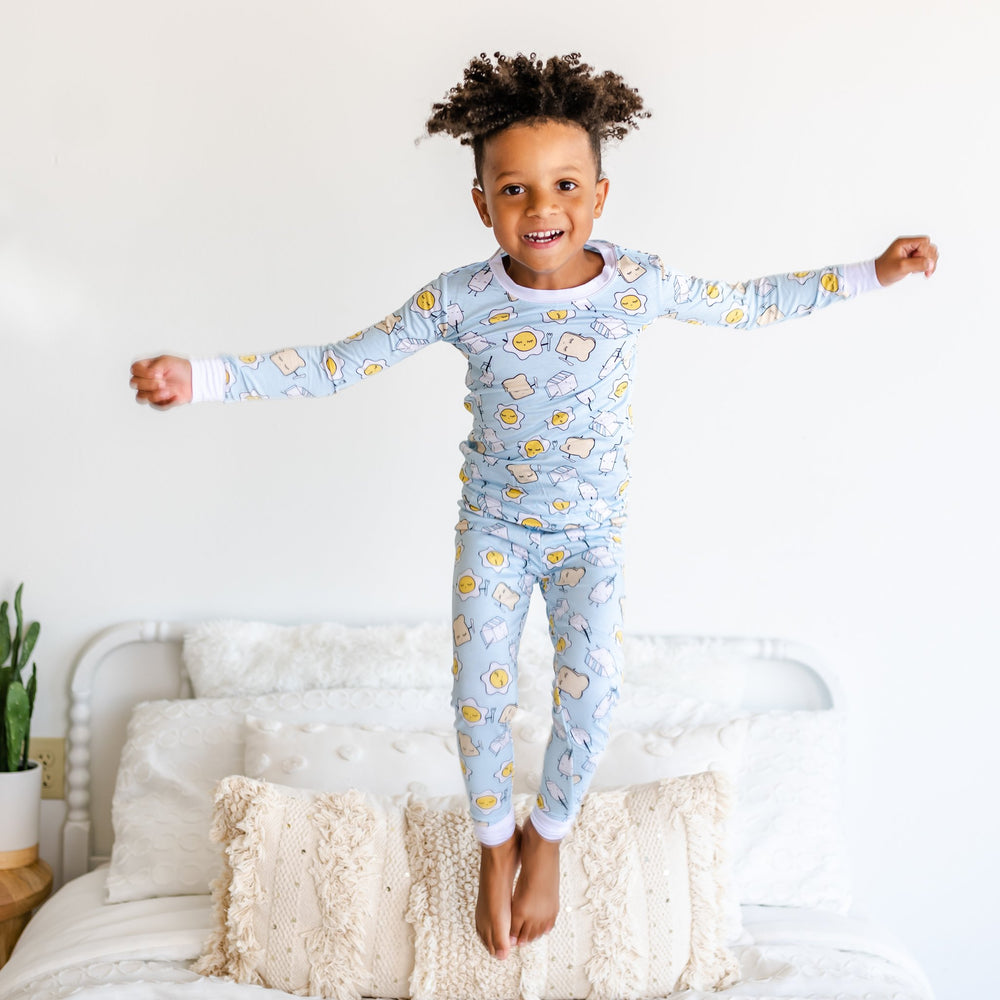 Image of toddler boy jumping on bed while wearing Blue Breakfast Buddies printed pajama set. This print has a light blue background with white trim accents and the breakfast foods featured on this print include sunny side up eggs, toast, and milk.
