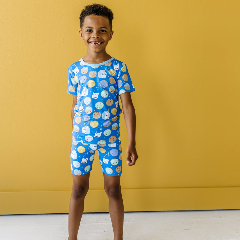 Image of little boy wearing a two-piece short sleeve and shorts pajama set in cookies and milk print. This print features milk cartons, colorful sprinkled cookies, and chocolate chip cookies that sit upon a blue background with sky blue trim.