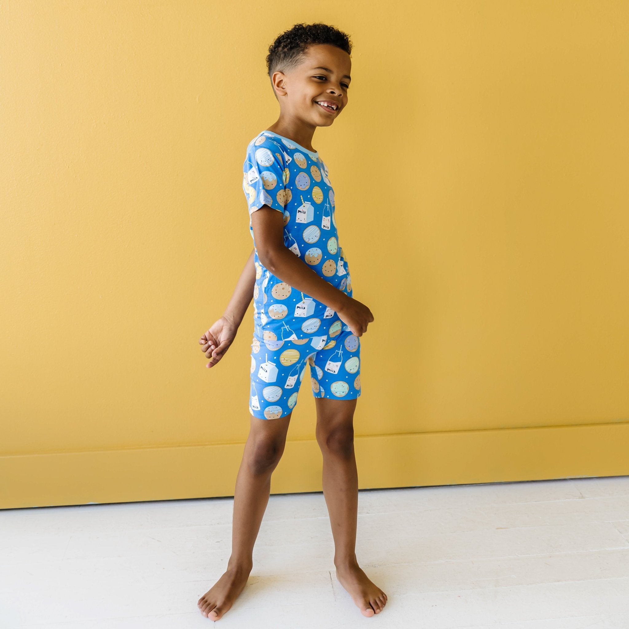 Image of little boy wearing a two-piece short sleeve and shorts pajama set in cookies and milk print. This print features milk cartons, colorful sprinkled cookies, and chocolate chip cookies that sit upon a blue background with sky blue trim.