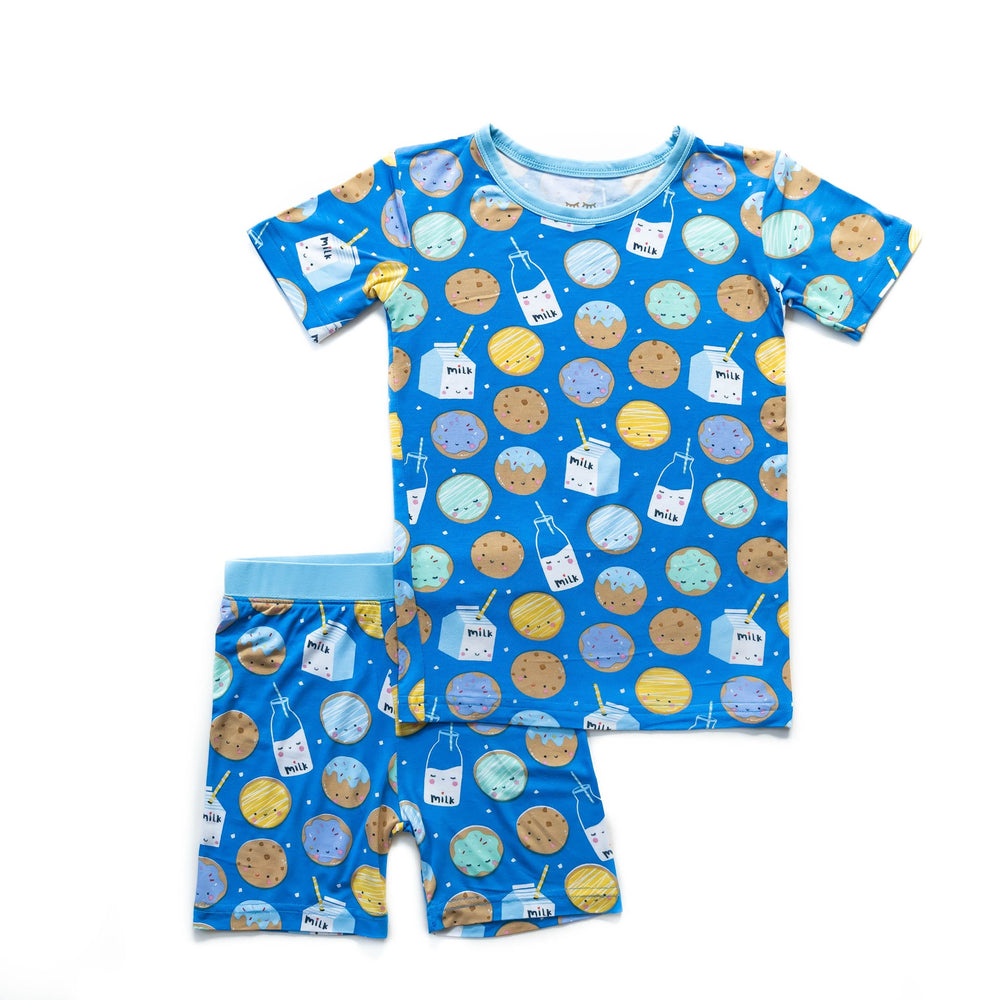 Flat lay image of short sleeve and shorts pajama set in cookies and milk print. This print features milk cartons, colorful sprinkled cookies, and chocolate chip cookies that sit upon a blue background with sky blue trim.