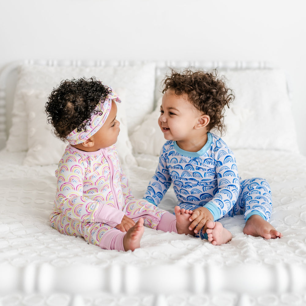Click to see full screen - image of baby girl and baby boy sitting on a bed with pink and blue rainbow printed pajamas. Baby boy is pictured wearing blue rainbow printed pajama set, which features a white background and shades of blue rainbows with sky blue trim accents. Baby girl is wearing pastel pink rainbow printed zip up romper with matching rainbow printed bow headband. 