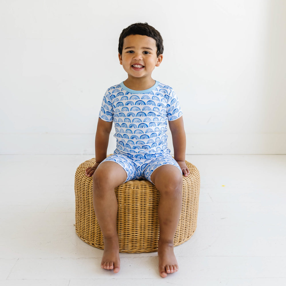 Click to see full screen - Image of toddler boy wearing a blue rainbow printed short sleeve and shorts pajama set. This print sits on a white background with shades of blue rainbows and sky blue trim details.