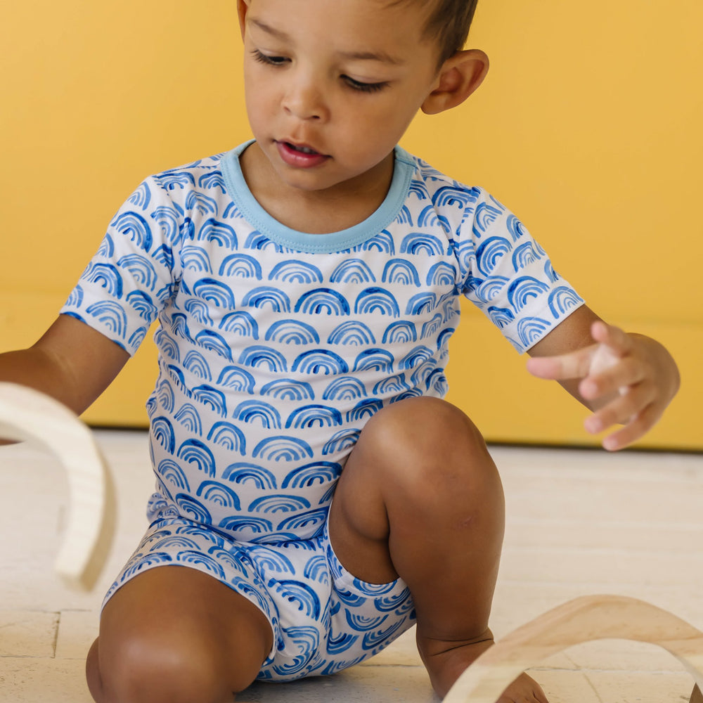 Image of toddler boy wearing a blue rainbow printed short sleeve and shorts pajama set. This print sits on a white background with shades of blue rainbows and sky blue trim details.