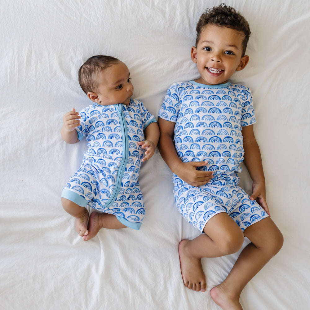 Image of two little boys lying next to each other, they are shown wearing matching blue rainbow printed pajamas. The infant boy is shown wearing a short sleeve and shorts zip up romper while the older boy is shown wearing a short sleeve and shorts pajama 
