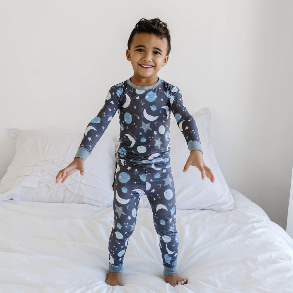 Image of little boy wearing a two-piece pajama set, featuring long sleeves and pants in blue to the moon and back print. This print features blue and gray moons, stars, and planets on a charcoal background with dusty gray trim.