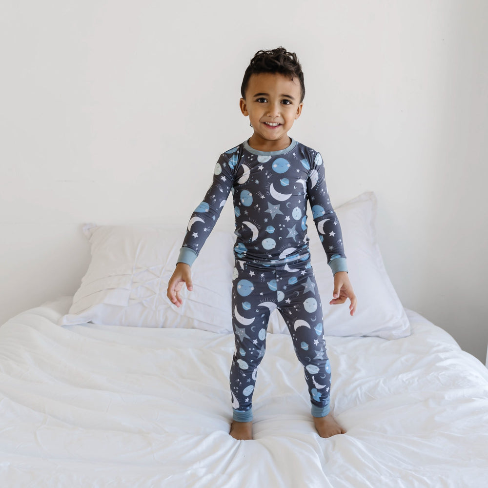 Image of little boy wearing a two-piece pajama set, featuring long sleeves and pants in blue to the moon and back print. This print features blue and gray moons, stars, and planets on a charcoal background with dusty gray trim.