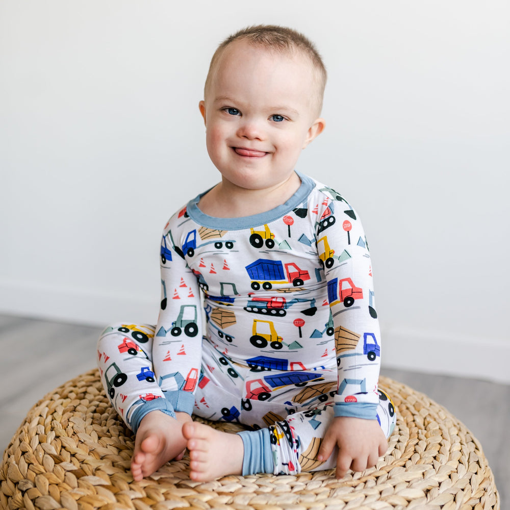 Image of little boy sitting on rattan pouf with construction printed pajamas. This print features utility trucks and tractors on a white background with sky blue trim accents.
