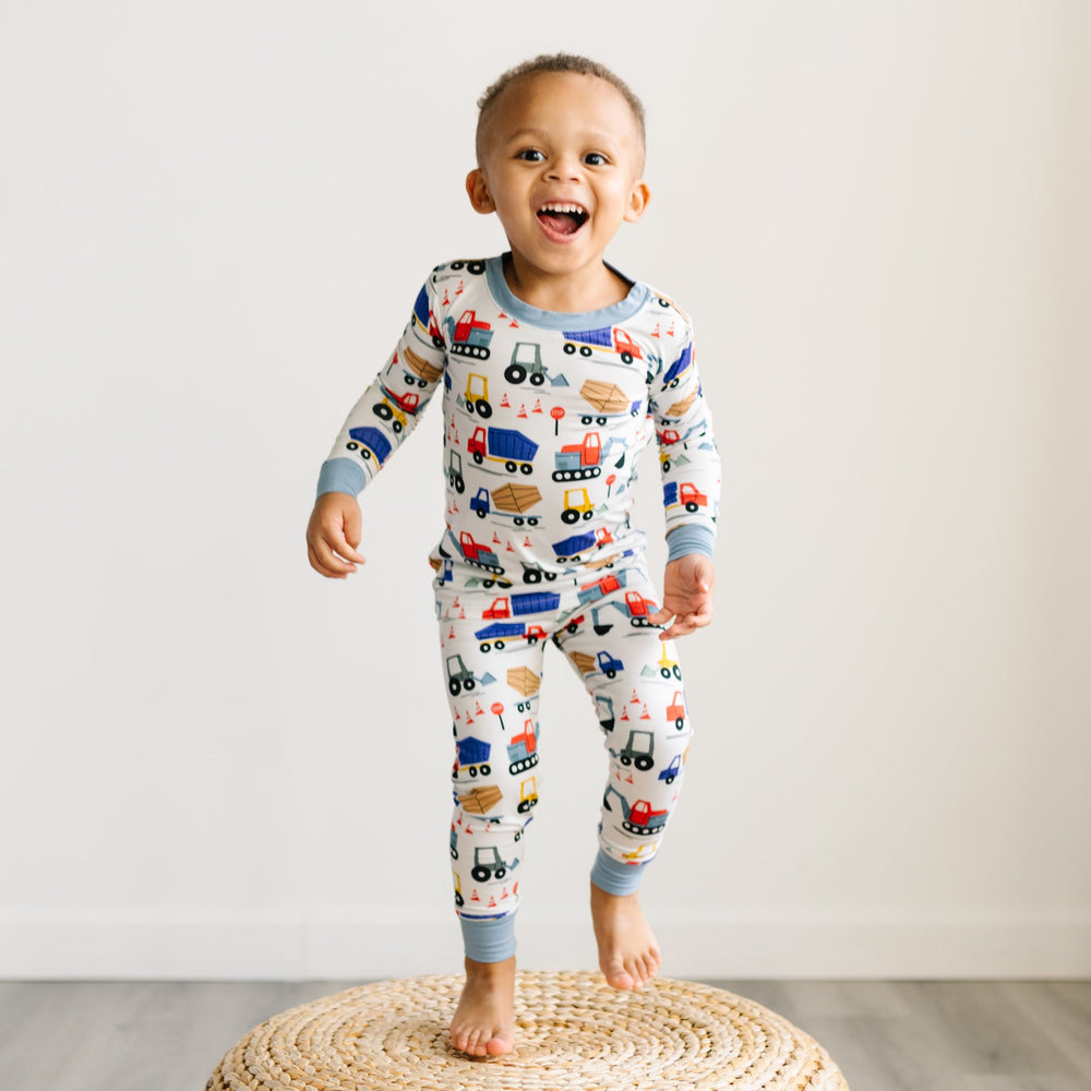 Click to see full screen - Image of toddler boy standing on rattan pouf. He is shown wearing two-piece pajama set in construction print. This print features utility trucks and tractors on a white background with sky blue trim accents.