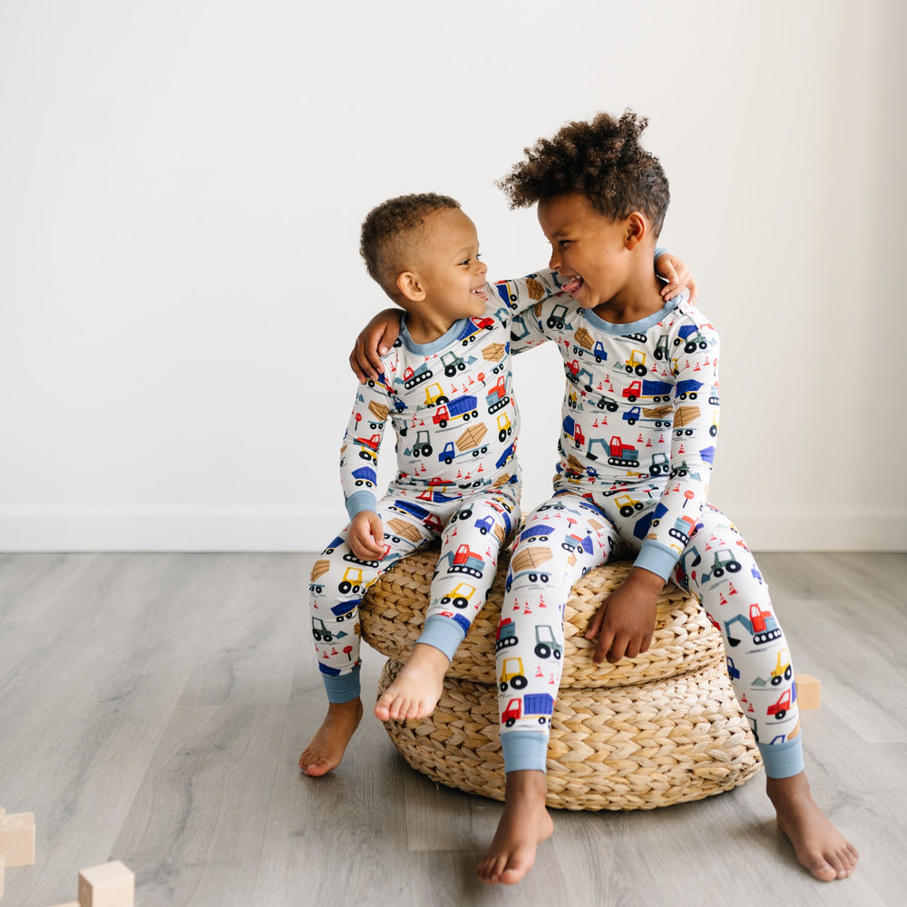Image of two boys sitting on rattan pouf wearing matching two-piece pajama sets in construction print. This print features utility trucks and tractors on a white background with sky blue trim accents.