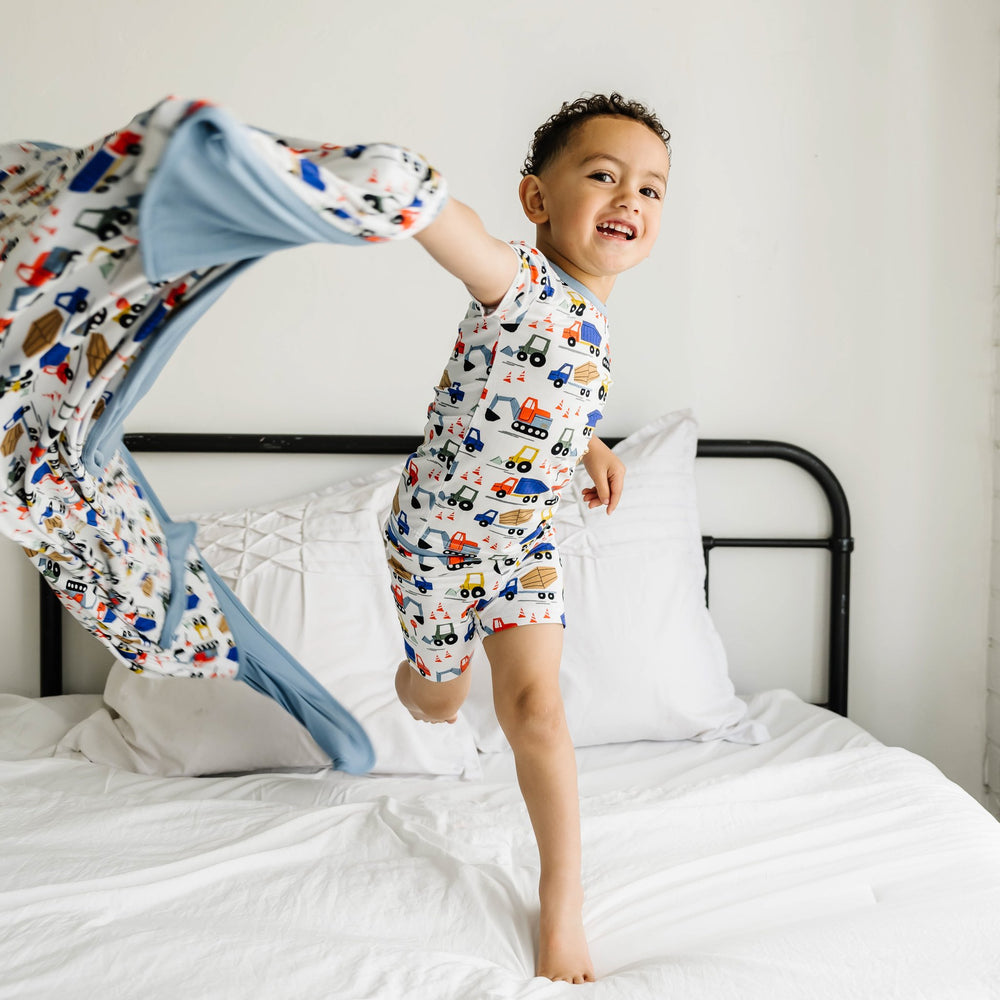 Image of infant boy holding a construction printed blanket. He is shown standing on a bed wearing a construction printed short sleeve and shorts pajama set. This print features utility trucks and tractors on a white background with sky blue trim accents.