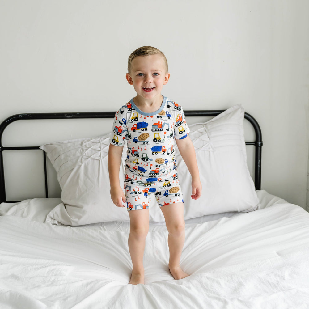 Image of toddler boy wearing a construction printed short sleeve and shorts pajama set. This print features utility trucks and tractors on a white background with sky blue trim accents.