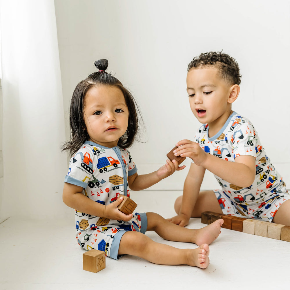 Image of two little boys playing with wooden building blocks. They are both shown wearing construction printed pajamas. One is shown wearing a short sleeve and shorts zip up romper and the other is shown wearing a short sleeve and shorts pajama set. Image of infant boy wearing a construction printed short sleeve and shorts zip up romper. This print features utility trucks and tractors on a white background with sky blue trim accents.