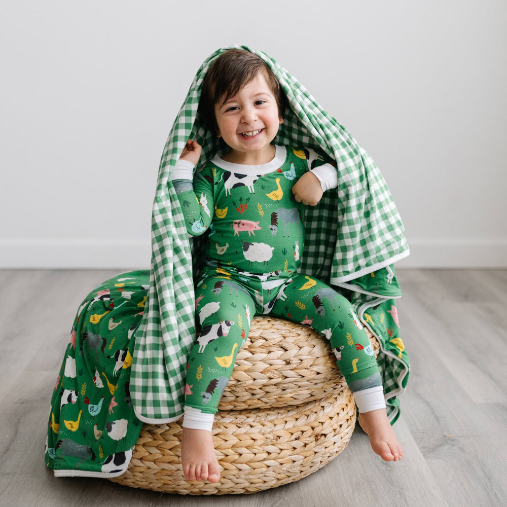 Image of toddler boy sitting on a rattan pouf with a green farm animals blanket draped around him. He is shown wearing a two piece pajama set in green farm animals print. The blanket features a double-sided mix and match print, with one side showing the g