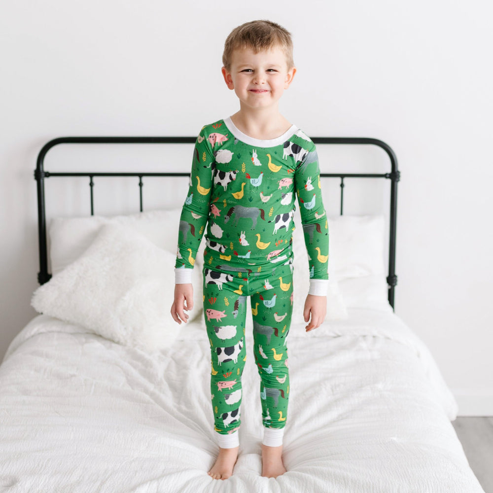 Image of little boy standing on a bed. He is shown wearing a two-piece pajama set in a green farm animals print. This print includes a green background with white trim details. The farm animals featured on this print include cows, pigs, ducks, sheep, pigs