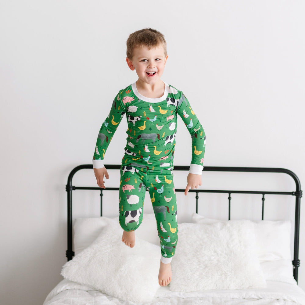 Image of little boy standing on a bed. He is shown wearing a two-piece pajama set in a green farm animals print. This print includes a green background with white trim details. The farm animals featured on this print include cows, pigs, ducks, sheep, pigs