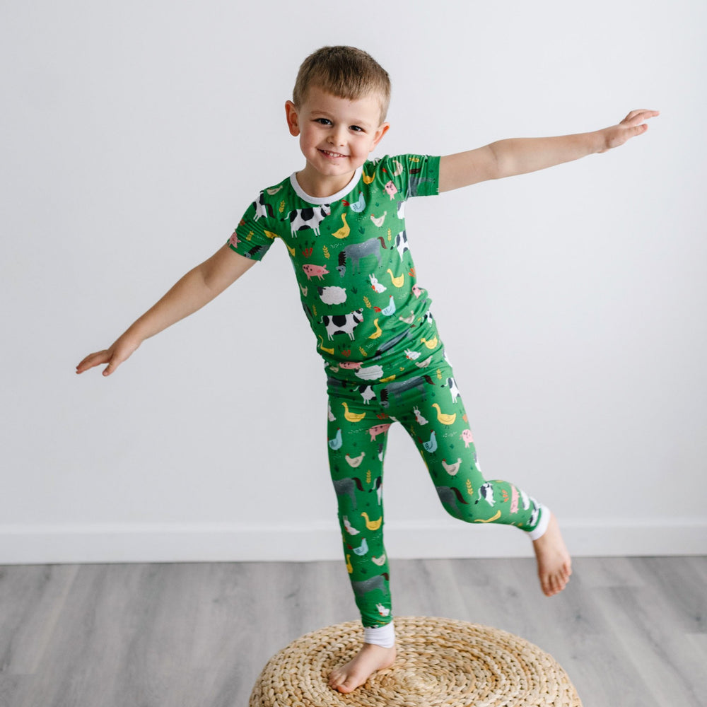 Image of a little boy standing on top of a rattan pouf. He is shown wearing a two-piece short sleeve pajama set in a green farm animals print. This print includes a green background with white trim details. The farm animals featured on this print include 