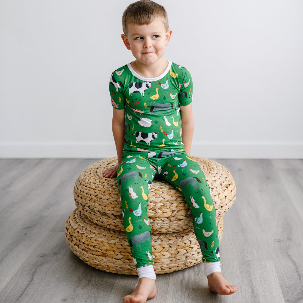 Image of a little boy sitting on a stack of rattan poufs. He is shown wearing a two-piece short sleeve pajama set in a green farm animals print. This print includes a green background with white trim details. The farm animals featured on this print includ