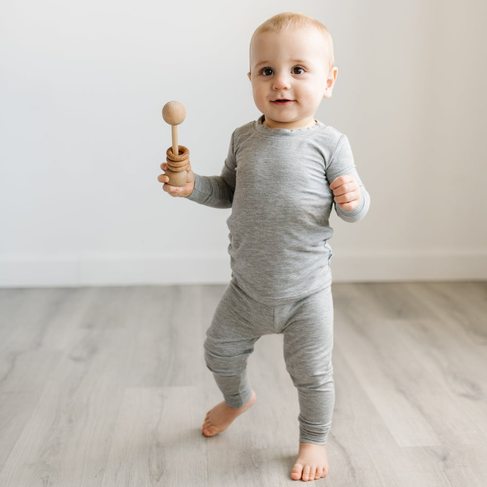 Image of toddler boy holding a wooden rattle. He is shown wearing two-piece pajama set in heather gray. 