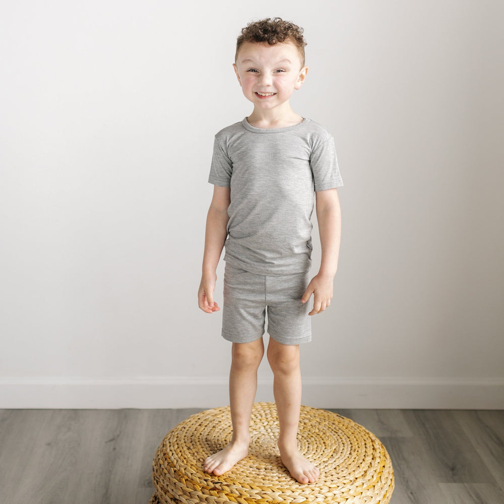 Image of little boy standing on a rattan pouf. He is shown wearing a two-piece short sleeve and shorts pajama set in heather gray.