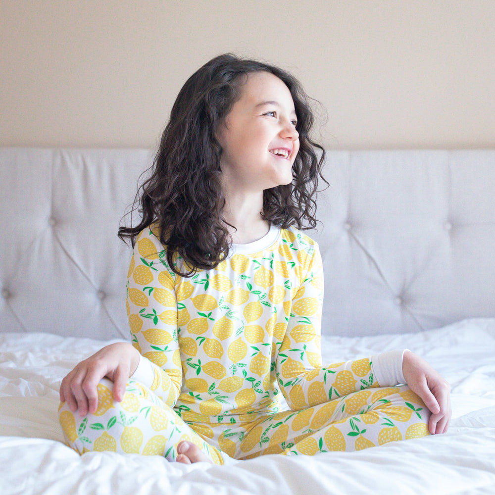 Image of little girl wearing Lemons printed two-piece pajama set. This print features vibrant pops of yellow fruit with green accents that sit upon a white background with white trim. 