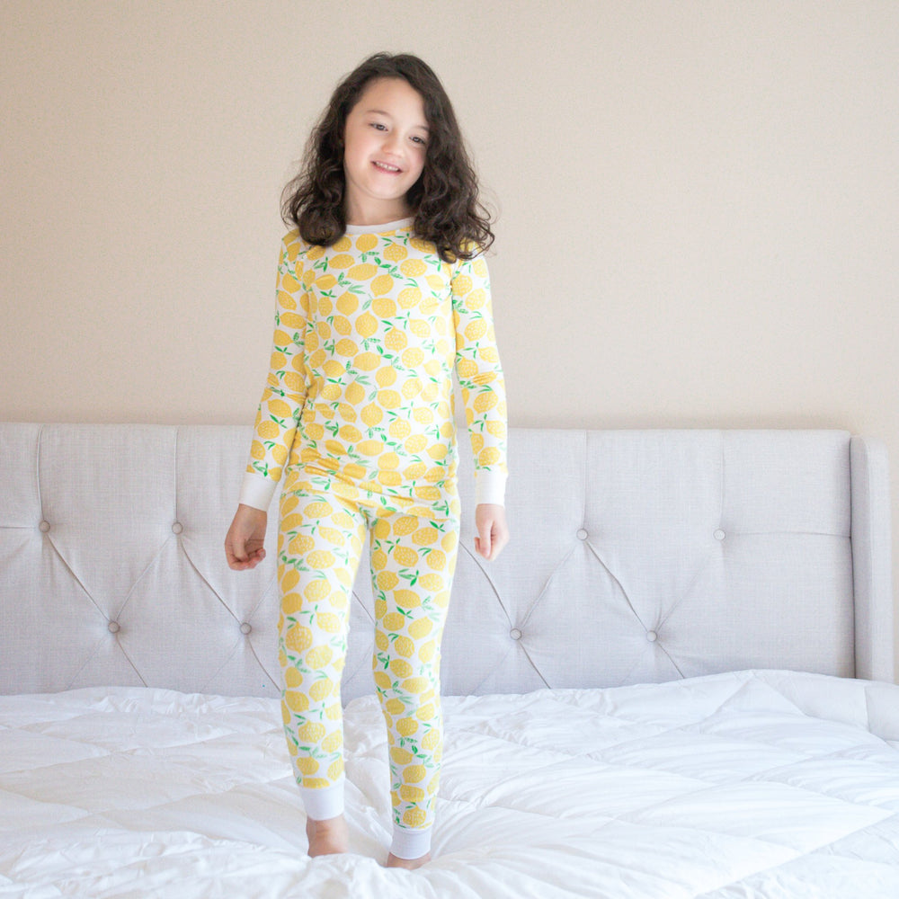 Image of little girl standing on the bed. She is shown wearing Lemons printed two-piece pajama set. This print features vibrant pops of yellow fruit with green accents that sit upon a white background with white trim. 