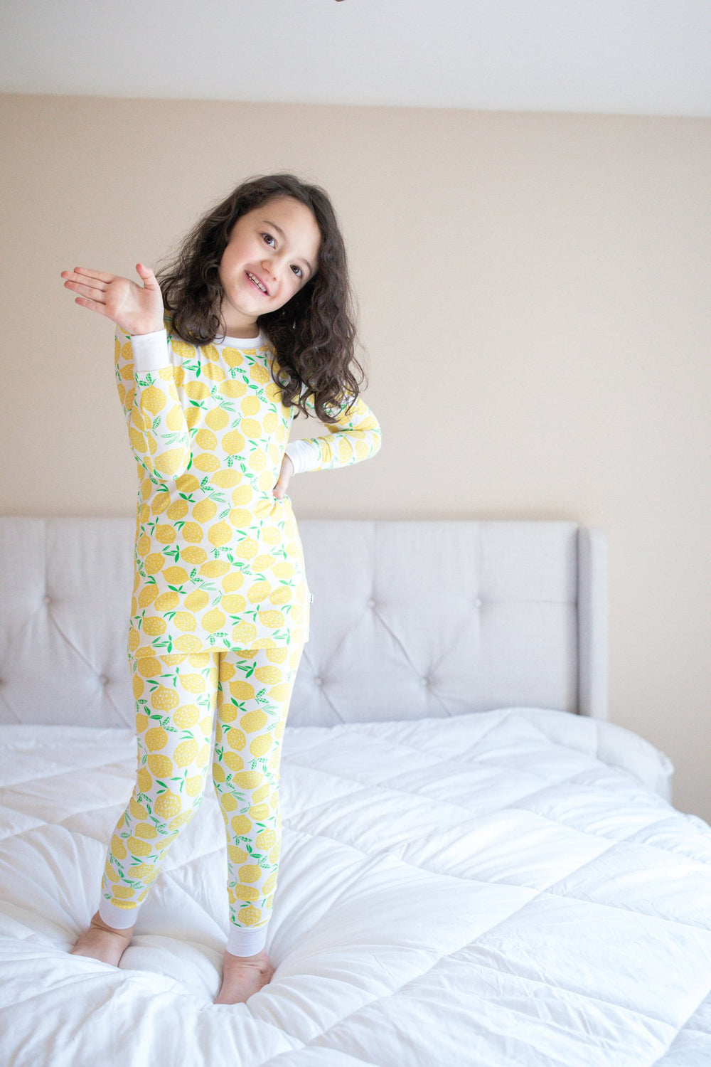 Image of little girl standing on the bed. She is wearing Lemons printed two-piece pajama set. This print features vibrant pops of yellow fruit with green accents that sit upon a white background with white trim. 